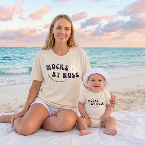 May include: A woman and a baby wearing matching beige t-shirts and a white hat, sitting on a white blanket on a beach. The shirts have the text "MOCKS BY ROSE" with a smiling face and a flower.