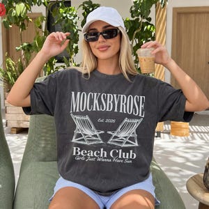 May include: A charcoal gray short-sleeve t-shirt featuring white screen-printed text that reads MOCKSBYROSE Est. 2026 Beach Club Girls Just Wanna Have Sun, accompanied by a graphic illustration of two beach lounge chairs.