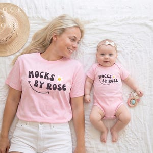 May include: A light pink t-shirt and baby onesie with the text "MOCKS BY ROSE" in black, a smiling face, and a daisy. The woman is wearing white shorts and a straw hat. The baby is holding a wooden toy.