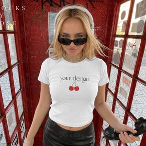 Könnte beinhalten: Ein weißes T-Shirt mit dem Text "your design" und einer Kirschgrafik. Das Model trägt eine Sonnenbrille und Kopfhörer und steht in einer roten Telefonzelle. Die Zelle hat Glasfenster und ein schwarzes Telefon.