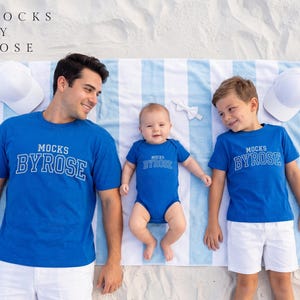 May include: Family matching blue t-shirts and a onesie with the text "MOCKS BY ROSE" in white. The family is lying on a striped blue and white towel on the beach. White baseball caps are placed on the sand.