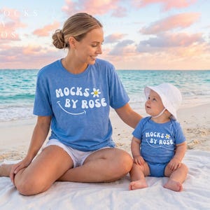 May include: A woman and a baby wearing matching blue t-shirts and sitting on a beach. The shirts have the text "MOCKS BY ROSE" with a daisy graphic. The baby wears a white sun hat. The background shows the ocean and a colorful sky.