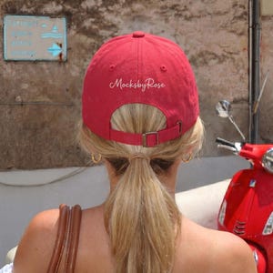 May include: A red baseball cap with the text "MocksbyRose" on the back. The cap has a curved brim and an adjustable strap. A red scooter is parked nearby.