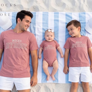 May include: A family wearing matching rose-colored t-shirts and a onesie with the text "MOCKS BY ROSE". The family is lying on a blue and white striped beach towel, with white hats on either side.