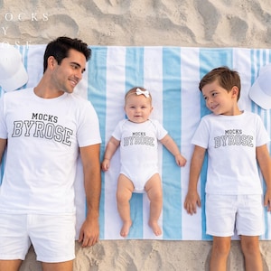 May include: A family of three wearing matching white t-shirts and shorts, and a baby wearing a white onesie, all lying on a striped blue and white beach towel. The shirts and onesie have the text "MOCKS BYROSE". Two white baseball caps are on the sand.