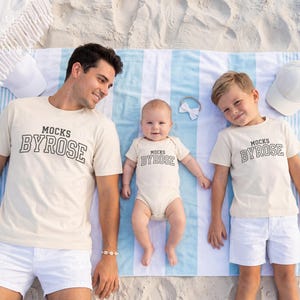 May include: A family of three, plus a baby, wearing matching beige t-shirts and a onesie with the text "Mocks Byrose" in black. They are lying on a striped blue and white beach towel, with white hats and a bow. The family is on a sandy beach.