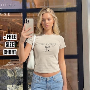 May include: A woman wearing a beige t-shirt with the text "your design" and a bow graphic. She is holding a phone and wearing blue jeans. The image also includes text that says "FREE SIZE CHART".