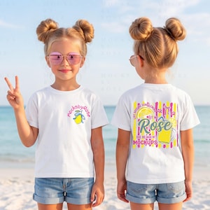 May include: Two white t-shirts with colorful graphics. One shirt has a small lemon design and the text "mocksbyrose". The other shirt has a lemon and drink graphic with the text "Rose" and "She be mixin' mockups". The shirts are worn by a child on a beach.