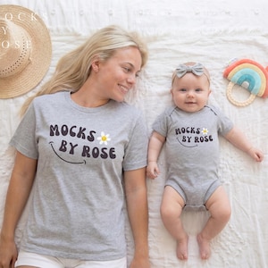 May include: A matching set of gray t-shirts and a baby onesie with the text "Mocks by Rose" and a daisy graphic. A straw hat and a rainbow-shaped baby toy are also visible. The items are on a white surface.