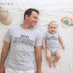 May include: A matching set of gray shirts and a baby onesie with the text "MOCKS BY ROSE". A beige baseball cap and a rainbow-colored baby toy are also visible. The items are on a white surface.