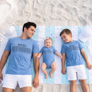 May include: A family of three wearing matching blue shirts and a baby bodysuit with the text "MOCKS BYROSE". They are lying on a striped beach towel with white shorts and a white hat. The scene is set on a sandy beach.