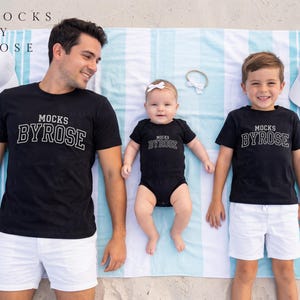 May include: A family of three wearing matching black t-shirts and a onesie with the text "MOCKS BYROSE" in white. They are lying on a striped blue and white beach towel, with white hats and a headband nearby. The theme is family apparel.