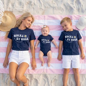 May include: A family of three wearing matching navy blue t-shirts and a onesie with the text "Mocks by Rose" and a daisy graphic. They are lying on a pink and white striped beach towel. A straw hat and white baseball cap are also visible.