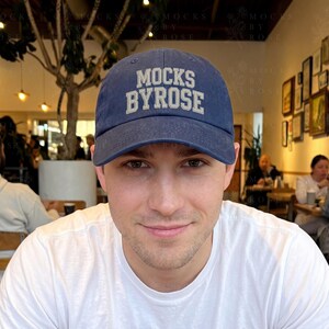 May include: A navy blue baseball cap with the text "MOCKS BYROSE" in white block letters. The cap is worn by a person in a white t-shirt. The background shows a restaurant setting with people and framed artwork on the wall.