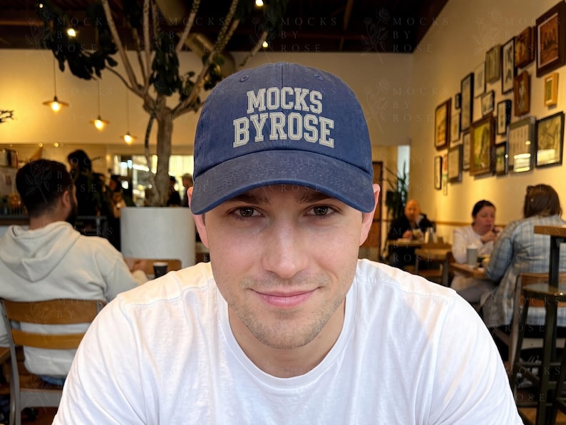 May include: A navy blue baseball cap with the text "MOCKS BYROSE" in white block letters. The cap is worn by a person in a white t-shirt. The background shows a restaurant setting with people and framed artwork on the wall.