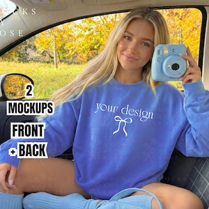 May include: A light blue sweatshirt with the text "your design" and a bow graphic. A light blue instant camera is held in the hand. The image also includes the text "2 Mockups Front + Back". The setting appears to be inside a car.