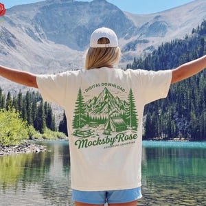 以下が含まれることがあります： 緑色の山とキャンプのシーンのグラフィックが特徴のクリーム色のTシャツ。「DIGITAL DOWNLOAD」と「Mocksby Rose Adventure Exporting」の文字が背面にプリントされています。山と湖の背景を背景に、屋外で着用されています。
