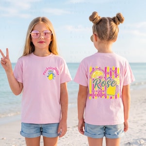 May include: Two young girls wearing pink t-shirts with graphic designs, standing on a beach. One shirt has a lemon graphic and the text "mocksbyrose". The other shirt has a lemon and text that says "Life gives you lemons but make Rose the best in mockups".