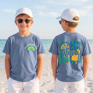 May include: Two children wearing blue t-shirts with a lime and lemon graphic. The front of the shirt has the text "mocksbyrose" and a lime graphic. The back of the shirt has a lemon and drink graphic with the text "Rose" and "Sip It Up".