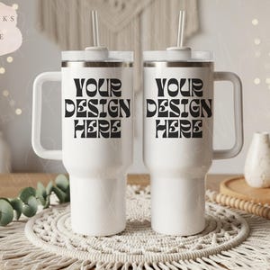 May include: Two white insulated tumblers with handles and lids, each with a straw. The tumblers have the text "YOUR DESIGN HERE" in black. The tumblers are on a woven placemat.