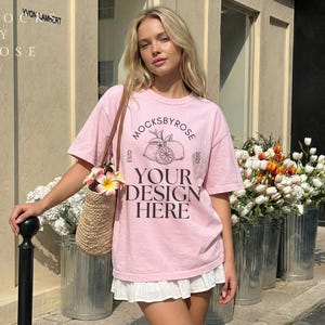 May include: A woman wearing a light pink t-shirt with a graphic design and the text "Mocksbyrose Your Design Here". She is carrying a straw bag with flowers. The image is taken outdoors with flowers in metal containers.