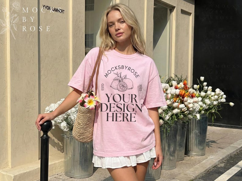 May include: A woman wearing a light pink t-shirt with a graphic design and the text "Mocksbyrose Your Design Here". She is carrying a straw bag with flowers. The image is taken outdoors with flowers in metal containers.