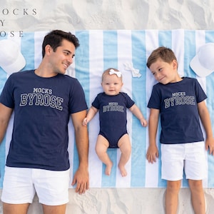 May include: A family wearing matching navy blue t-shirts and a onesie with the text "MOCKS BY ROSE" in white. They are lying on a blue and white striped beach towel with white hats. The background is sand.
