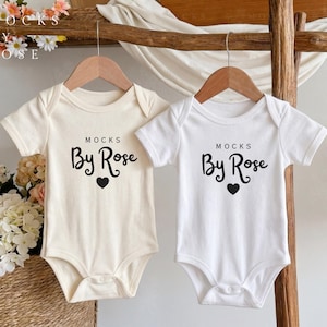 May include: Two baby onesies, one cream and one white, hang on wooden hangers. Each onesie has the text "MOCKS By Rose" in black script with a black heart below. The background includes flowers and a wooden ladder.