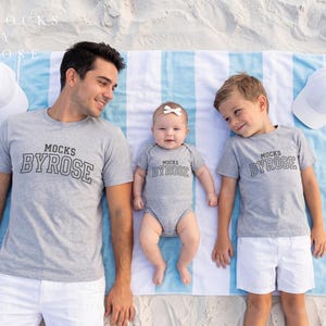 May include: A family wearing matching gray t-shirts and a onesie with the text "MOCKS BYROSE" in a collegiate font. The family is lying on a blue and white striped beach towel with two white baseball caps.