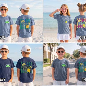 May include: Multiple children's t-shirts in various colors, including blue jean, true navy, and denim. Each shirt features a front pocket design and a larger graphic on the back with text and illustrations. The shirts are displayed on children in front of beach and street backgrounds.
