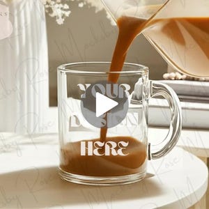 May include: A clear glass mug with a handle is being filled with a brown liquid. The mug has the text "YOUR DESIGN HERE" printed on it. The scene is set on a white surface with a blurred background.