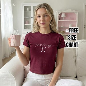 Può includere: Una donna indossa una maglietta bordeaux con la scritta "your design" in bianco. Tiene in mano una tazza rosa con un fiocco. L'immagine include una grafica "+ FREE SIZE CHART".