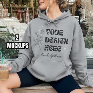May include: A grey hoodie featuring the text "YOUR DESIGN HERE" and a floral illustration. The hoodie has a front pocket and a drawstring hood. The image also includes the text "MocksbyRose" and "2 MOCKUPS". The person is holding a clear plastic cup with a straw.