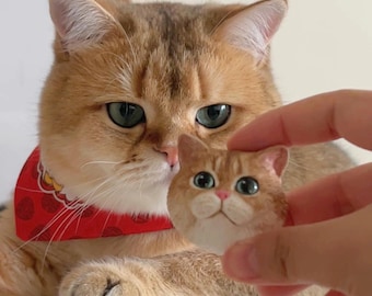 Könnte beinhalten: Eine goldene Tabby-Katze mit einem roten Bandana mit weißem und rotem Muster schaut auf eine kleine, handbemalte Keramik-Katzenfigur, die in einer Hand gehalten wird.