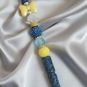 May include: A decorative pen with a blue, yellow, and silver design. The pen features a bow, crown, and beaded accents. The pen's barrel is covered in blue rhinestones, and the tip is silver.