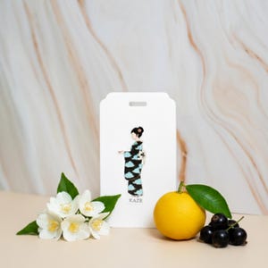 May include: A white tag with a drawing of a woman in a kimono with black and blue cloud patterns. The tag has the word "KAZE" printed on it. The tag is next to a yellow lemon, white flowers, and dark berries.