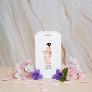 May include: A white tag featuring a woman in a kimono, with the word "SAXURA" below. The tag is surrounded by pink and purple flowers and petals. The background has a marble pattern.