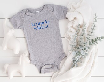 May include: A gray baby onesie with the text "kentucky wildcat" in blue. The onesie is laid on a white wooden surface next to a white blanket, a decorative elephant, and a unicorn figurine.