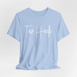May include: A light blue t-shirt with the words "Tar Heels" in white script across the front. The shirt has short sleeves and a crew neck. The fabric is slightly wrinkled.