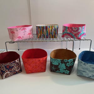 May include: Seven fabric storage bins with different patterns. The bins are lined up on a metal shelf. The patterns include flamingos, paisley, red floral, turquoise floral, blue tie-dye, and rainbow stripes.