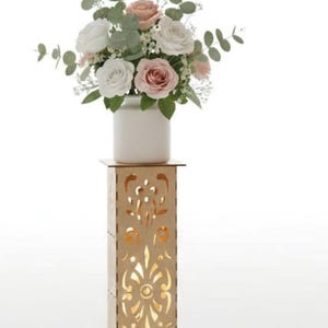 Puede incluir: Un arreglo floral en un jarrón blanco se encuentra sobre un soporte de madera decorativo con diseños calados. El arreglo incluye rosas blancas y rosas con vegetación. El texto "INCLUDES 10 SIZES" está en letras doradas.