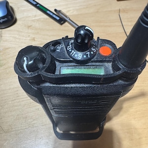 Pode incluir: Um rádio bidirecional preto Motorola APX 6000. O rádio tem um visor verde, vários botões e uma antena. Um botão laranja é visível na parte superior. O rádio está sobre uma superfície de madeira marrom clara.