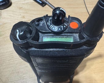 May include: A black Motorola APX 6000 series two-way radio. The radio has a green display, several knobs, and an antenna. An orange button is visible on the top. The radio is on a light brown wooden surface.