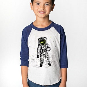 Peut inclure: Un t-shirt de baseball blanc et bleu avec un graphique d'un astronaute sur la lune. L'astronaute porte une combinaison spatiale et un casque. Le t-shirt a une manche bleu marine et une manche blanche.