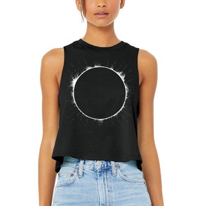 Woman is wearing a black, racerback cropped tank top printed with a total solar eclipse design on the front in white ink. Shirt by Little Lark