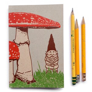 May include: A brown paper notebook with a red and white mushroom and a brown gnome illustration. The notebook is next to three yellow pencils.