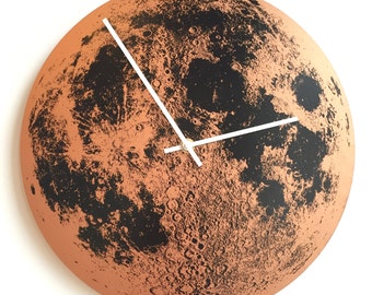May include: A round wall clock with a terracotta-colored face, featuring a detailed black lunar surface design. White clock hands indicate the time. The clock's diameter is approximately 12 inches.