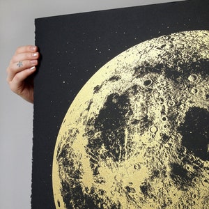 May include: A black and gold print of the moon with a detailed depiction of its surface. The moon is surrounded by a black background with small white stars.