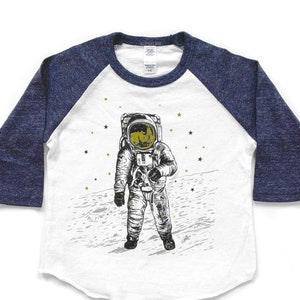 Peut inclure: Un t-shirt de baseball blanc et bleu avec un graphique d'un astronaute debout sur la lune. L'astronaute porte une combinaison spatiale blanche avec un casque dor&eacute; et est entour&eacute; d'&eacute;toiles dor&eacute;es.