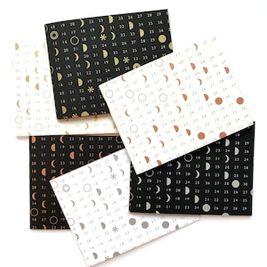 May include: A stack of six small notebooks with different colored covers. Each cover features a repeating pattern of gold, silver, or copper moons and stars on a black, white, or gray background. The numbers 14 through 30 are printed on each cover.